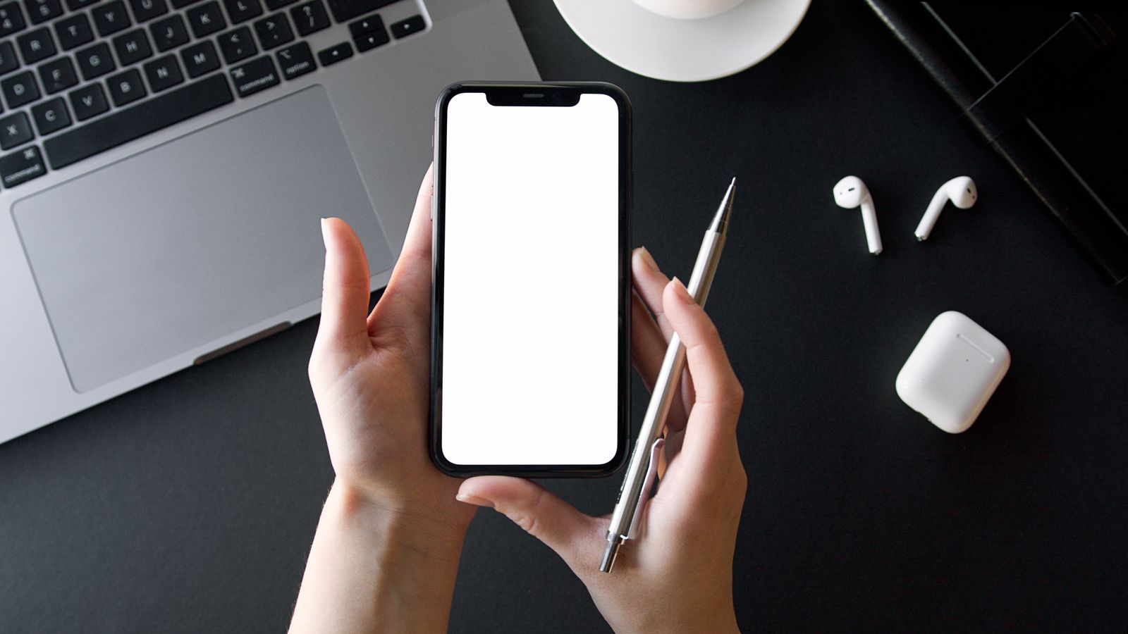 Hands holding a blank-screen phone and pen beside a laptop